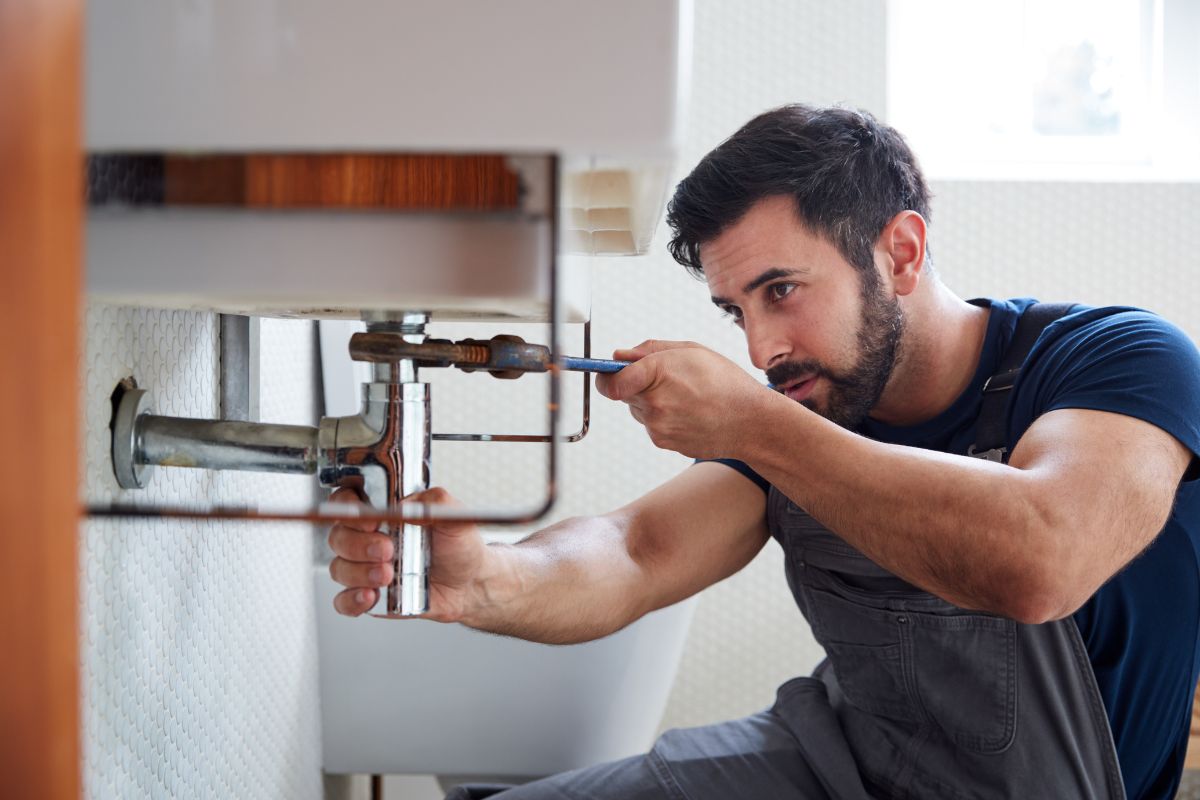 A plumber in a blue shirt and work apron tightens the pipes under a white sink.