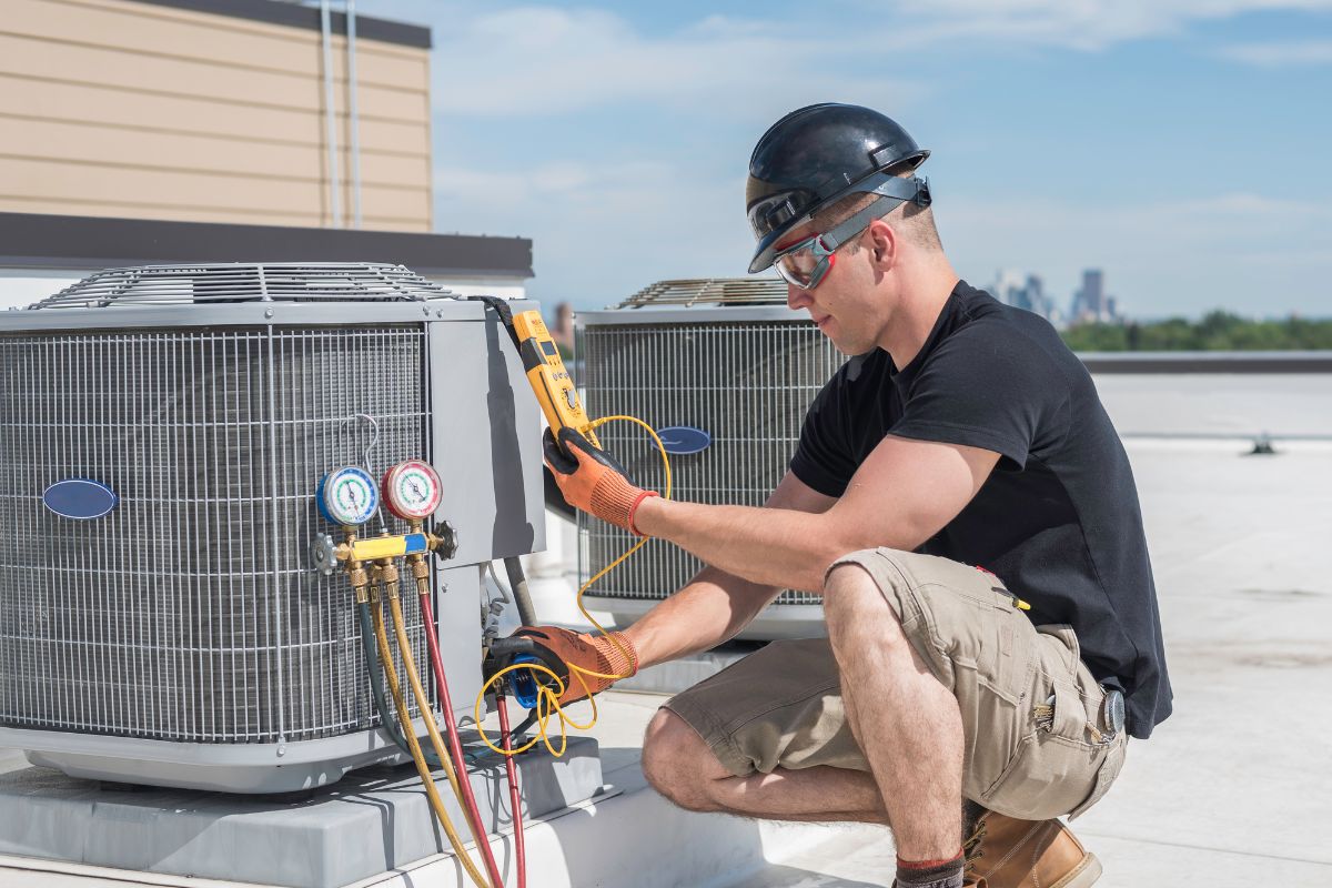 An HVAC contractor in a black shirt and shorts wears a work helmet - reading a meter next to an air conditioner.