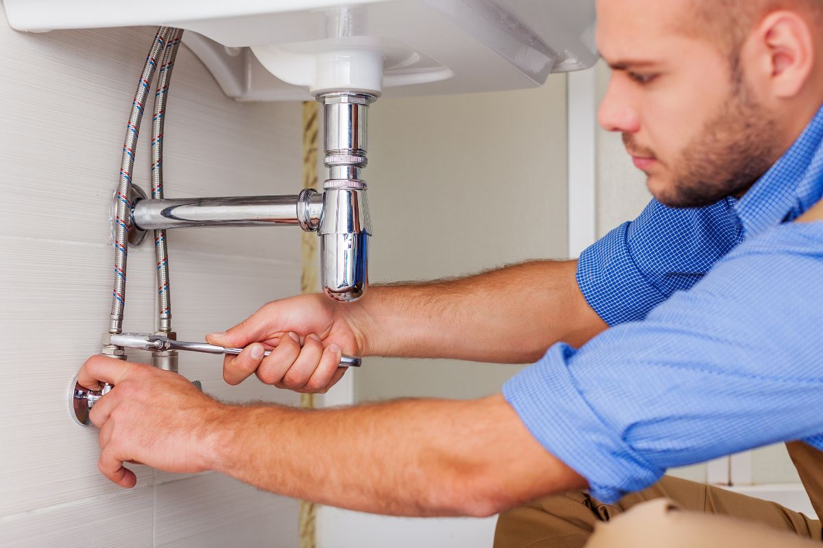 A close-up of a plumber in a blue shirt tightening a sing pipe with a wrench.